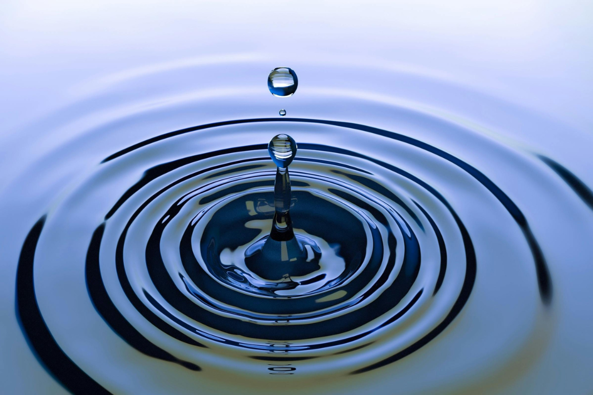 Close-up of a water droplet falling into a calm surface, creating circular ripples in blue-toned light.